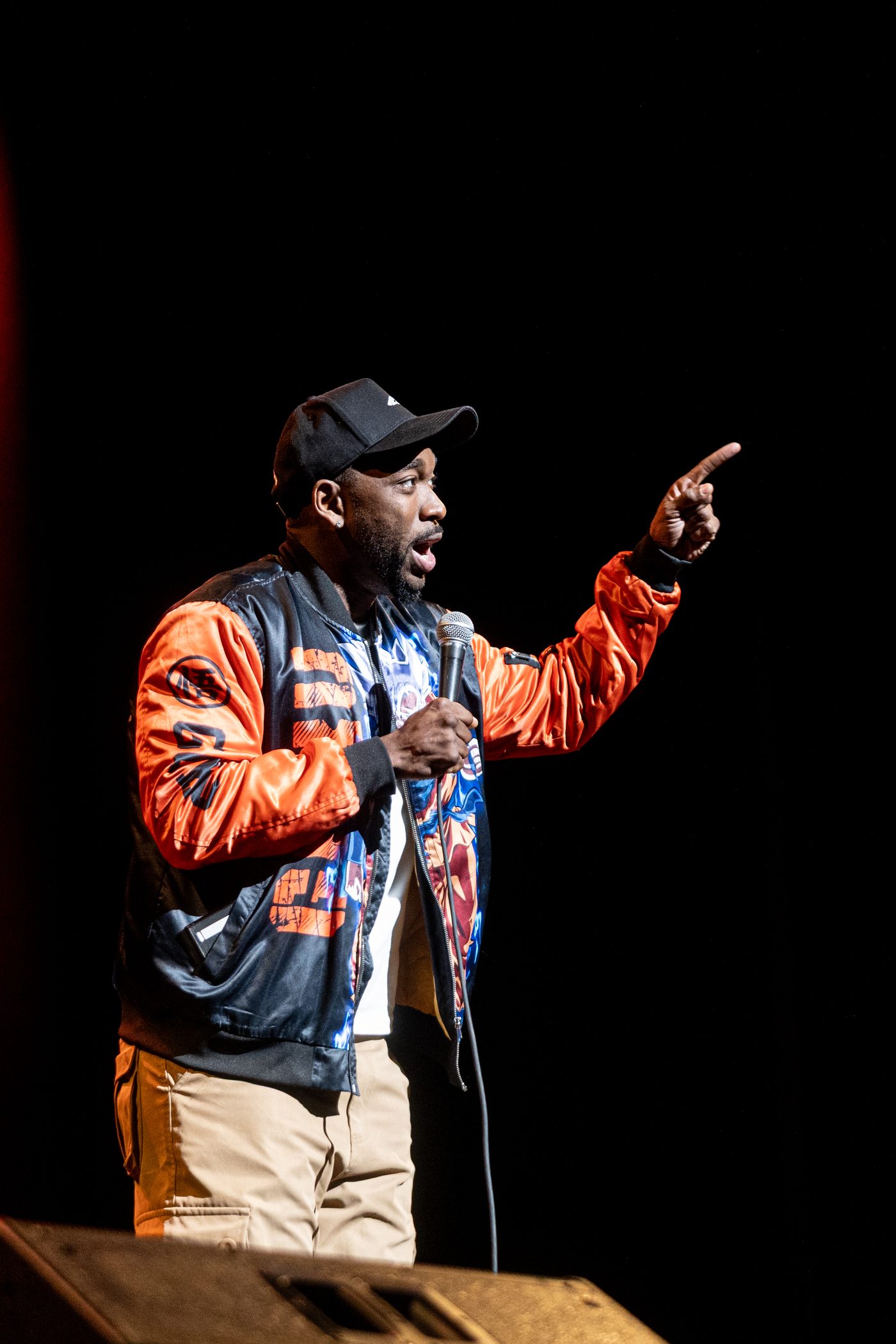 Stand-up comedian mid-set on a darkened stage, pointing toward the audience