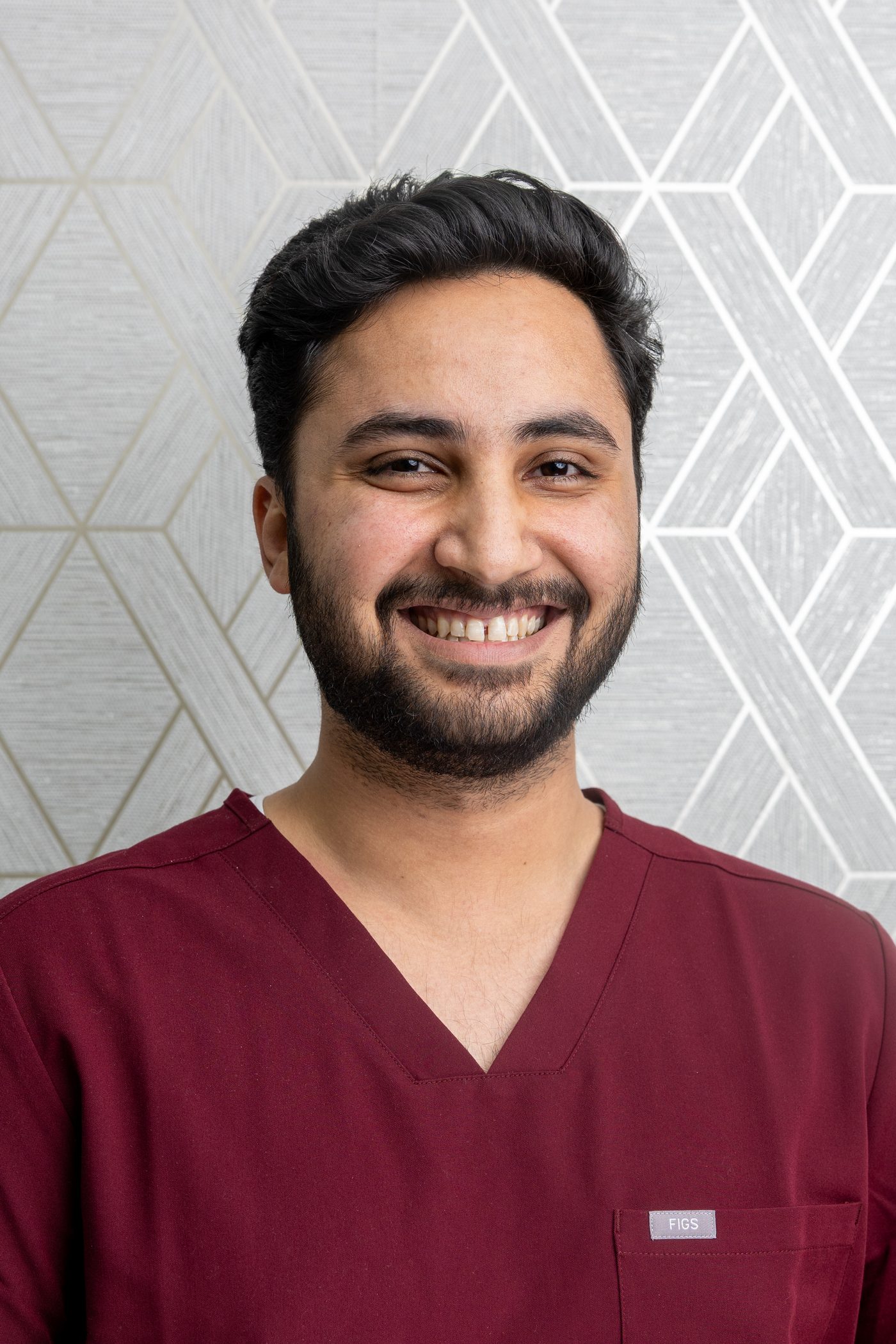 Studio headshot of a smiling healthcare professional in maroon scrubs against a patterned wall
